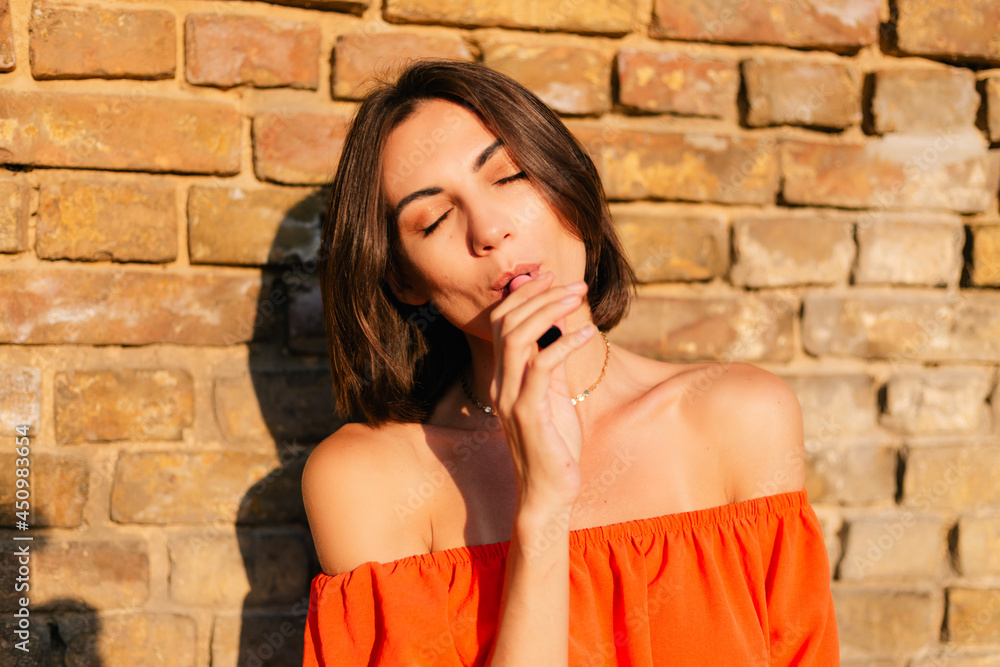 Stylish woman in orange clothes at sunset at brick wall background smoking electronic cigarette Stylish woman in orange clothes at sunset at brick wall background smoking electronic cigarette