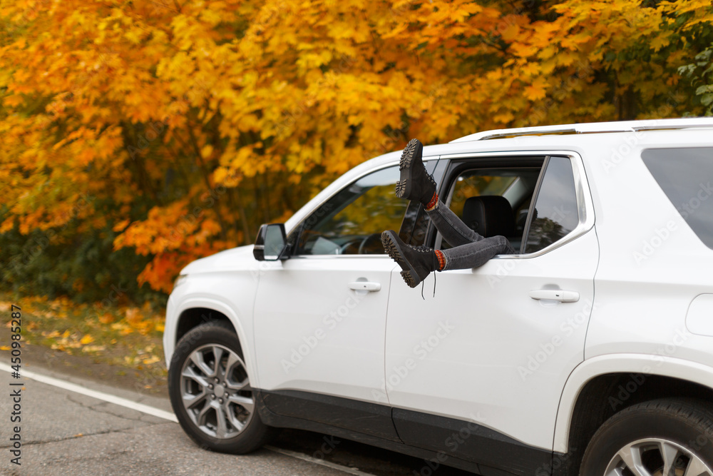 Stylish unrecognizable woman in grey jeans and black boots put feet out of white car window. Yellow colorful maple leaves on background. Freedom and relaxation on road trip by car. Traveller lifestyle Stylish unrecognizable woman in grey jeans and black boots put feet out of white car window. Yellow colorful maple leaves on background. Freedom and relaxation on road trip by car. Traveller lifestyle