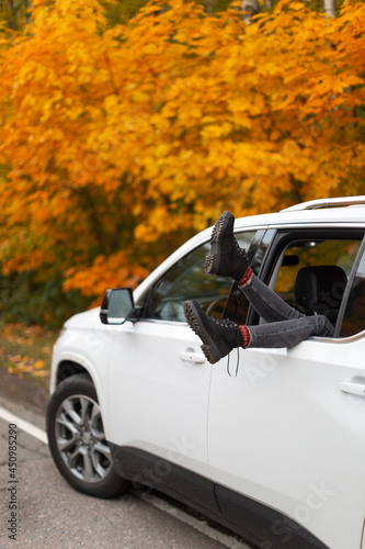 Unrecognizable woman in grey jeans and stylish black boots put feet out of white car window. Yellow colorful maple leaves on background. Traveller lifestyle. Selective focus  vertical photo  close up.