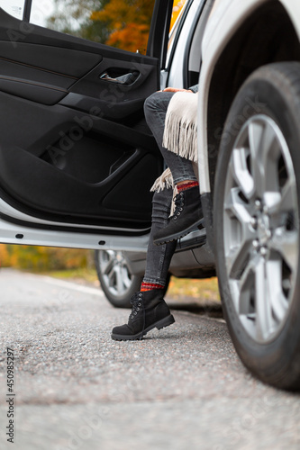 Stylish unrecognizable girl in grey jeans, beige scarf and black boots sitting in car doorway. One foot staying on asphalt. Selective focus on leg. Autumn season, colorful background. Roadtrip concept © Denis Mamin