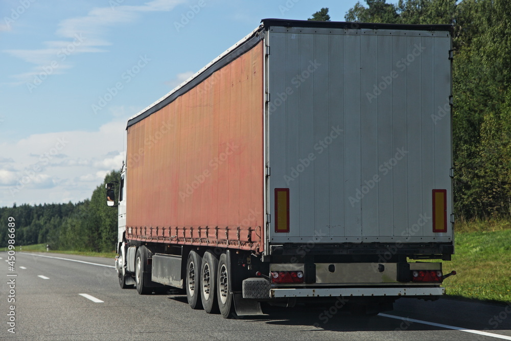 One European brown semi trucks back side view on free suburban highway road at Sunny summer day on green forest background, cargo transportation logistics traffic landscape