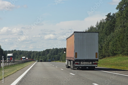 One European brown semi trucks back side view on empty suburban highway road at Sunny summer day on green forest background, cargo transportation logistics traffic landscape