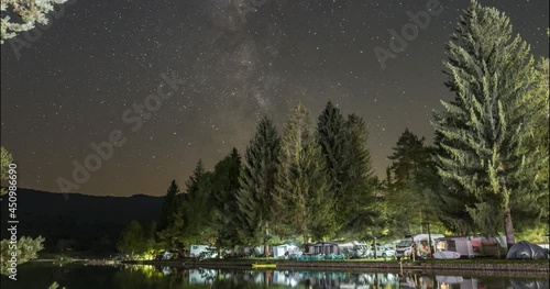 Night time lapse of the milky way galaxy and stars moving over Sobec campground in summer. Camp and small pond in Slovenia. Silhouette of mountain range. Static shot, wide angle photo