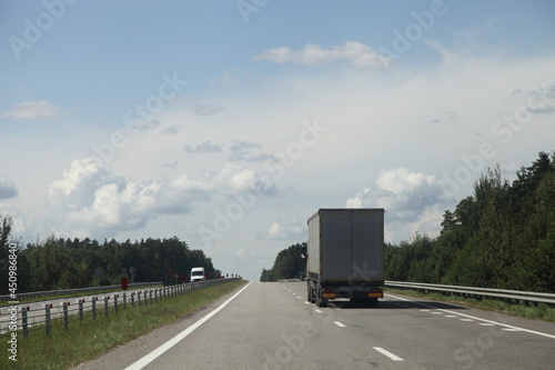 European heavy truck move on the countryside highway road at Sunny summer day on green forest, road fence and blue sky with clouds background, back side view, transportation logistics © Ilya
