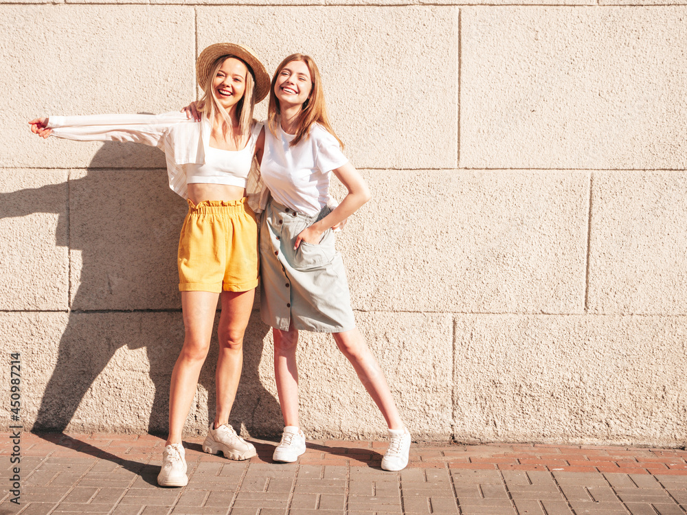 Two young beautiful smiling hipster female in trendy summer clothes.Sexy carefree women posing near white wall in the street. Positive pure models having fun at sunset, hugging and going crazy