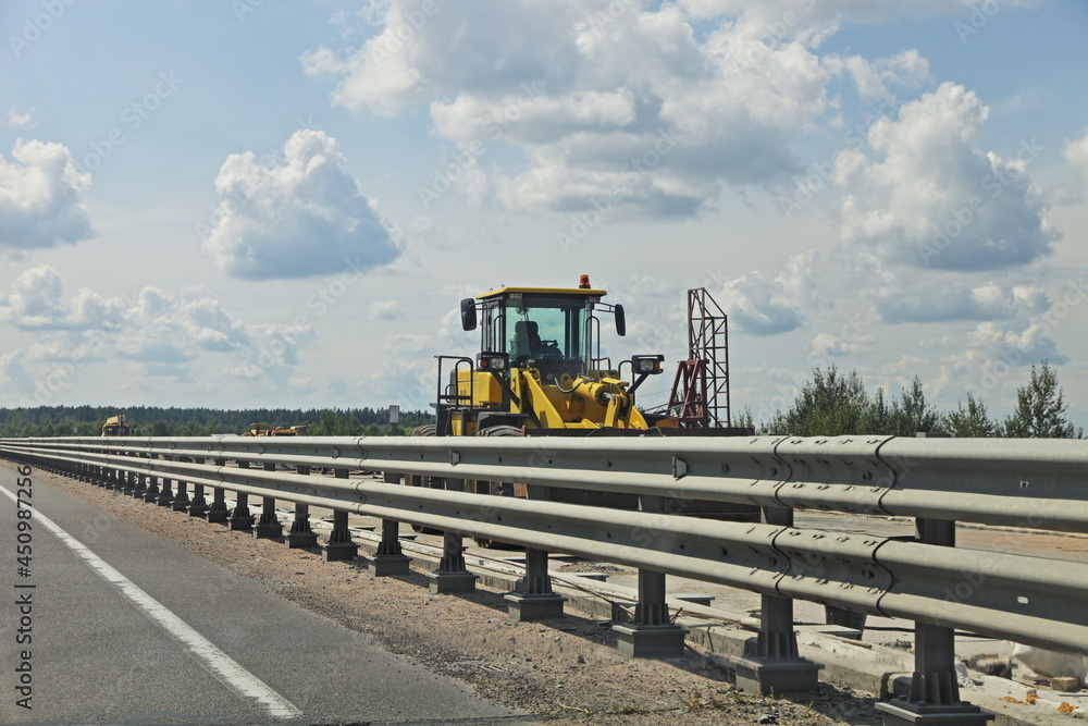 Yellow bulldozer wheeled tractor on highway fence and blue sky with clouds background at Sunny summer day , road bridge repair and building landscape Yellow bulldozer wheeled tractor on highway fence and blue sky with clouds background at Sunny summer day , road bridge repair and building landscape