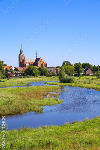 View over green wetland water on cityscape of durch town with tower of medieval church against blue summer sky - Neer (Limburg), Netherlands
