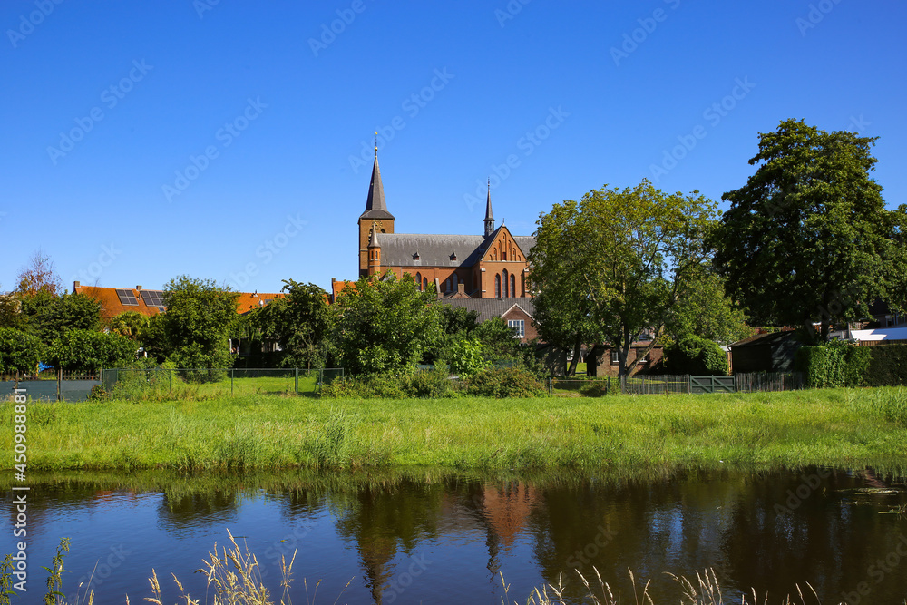 View over river and green pasture on small dutch idyllic village with medieval old church against blue cloudless summer sky - Neer (Limburg), Netherlands