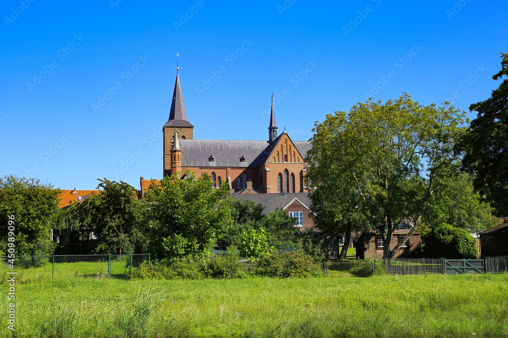 View over lush green pasture and trees on small dutch idyllic village with medieval old church against blue cloudless summer sky - Neer (Limburg), Netherlands