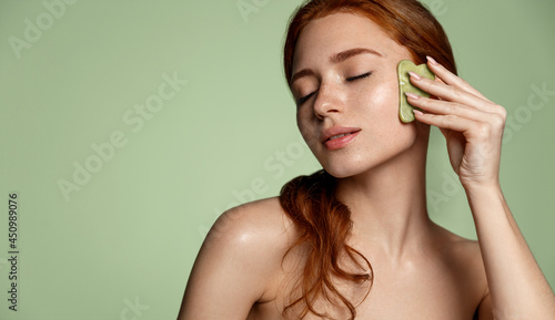 Beautiful redhead woman doing skin care routine after shower, scrapping, massage facial skin with jade gua sha scrapper, body and face beauty concept, green background photo