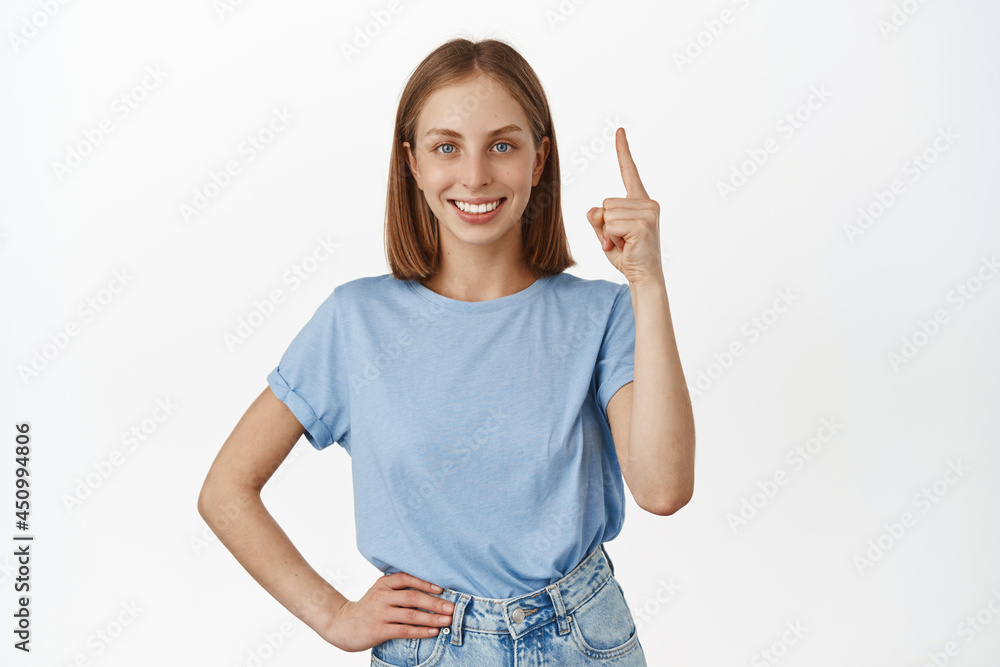 Portrait of attractive young woman with blond hair and blue eyes, pointing finger up at promo, showing sale dicount banner and smiling satisfied, white background Portrait of attractive young woman with blond hair and blue eyes, pointing finger up at promo, showing sale dicount banner and smiling satisfied, white background