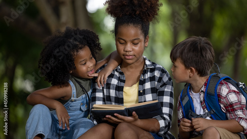 Sister black skin African American ethnicity sitting at on tree base nature reading to her younger siblings a book about adventure stories