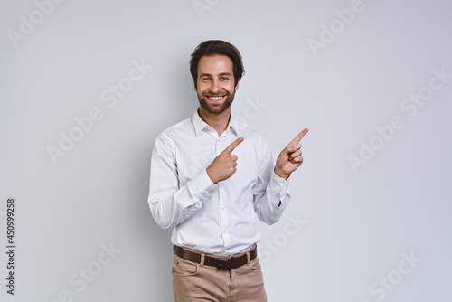 Handsome young smiling man in white shirt looking at camera and pointing away while standing against gray background