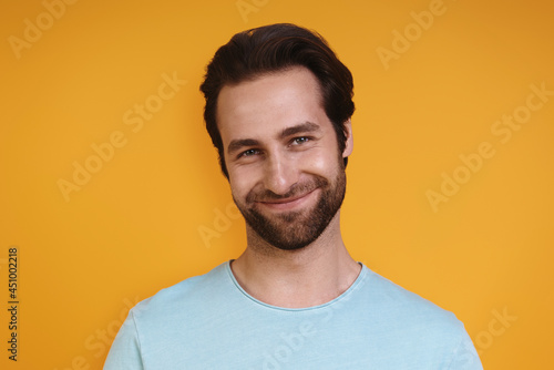 Portrait of young man in casual clothing looking at camera and smiling while standing against yellow background © gstockstudio