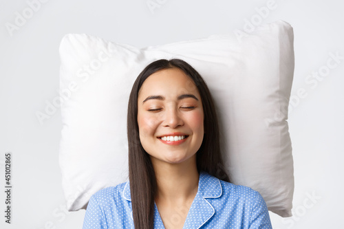 Smiling dreamy and beautiful asian female lying in bed on pillow, wearing blue pajamas, close eyes and grinning, daydreaming or sleeping at night, imaging something cute, white background photo