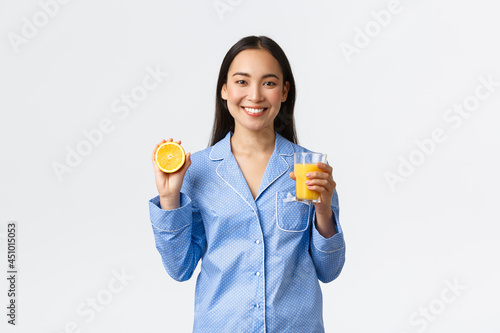 Morning, active and healthy lifestyle and home concept. Smiling cheerful asian girl starting her day with fresh made orange guice, holding glass and half of orange, looking happy and energized photo