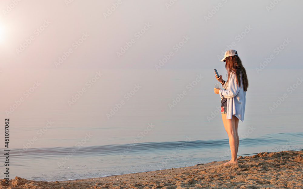 Girl taking a photo of sunrise on smartphone on the seashore in morning sun rays.
