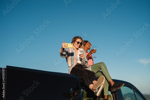 Diverse friends having fun doing video call party celebrating with wine while sitting on top of camper - Focus on left woman face