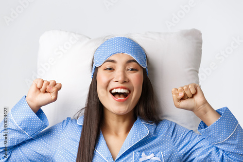 Close-up of enthusiastic asian girl in blue pajamas and sleeping mask, stretching hands up delighted after good night sleep, take-off eyemask in morning, lying in bed on pillow and smiling happy photo