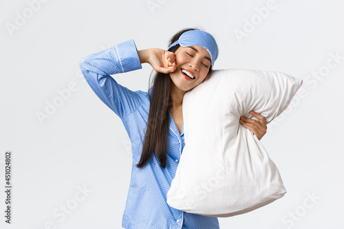 Smiling pleased cute asian girl in blue pyjama and sleeping mask, hugging pillow and stretching hands delighted as finally going bed, want sleep or waking up in morning, white background photo