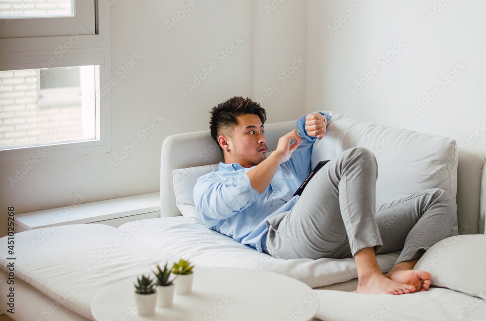 Young handsome Chinese man at home sitting on couch using digital tablet