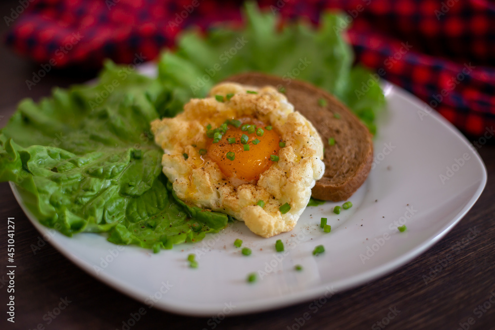 Fried egg on a white plate with leaves of salad and bread, brown table and red textile