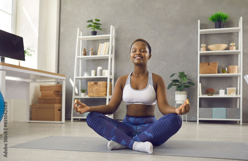 Happy relaxed short haired young black woman in modern sportswear sitting in cross legged yoga pose on floor at home, smiling eyes closed, enjoying meditation and filling herself with positive energy Happy relaxed short haired young black woman in modern sportswear sitting in cross legged yoga pose on floor at home, smiling eyes closed, enjoying meditation and filling herself with positive energy
