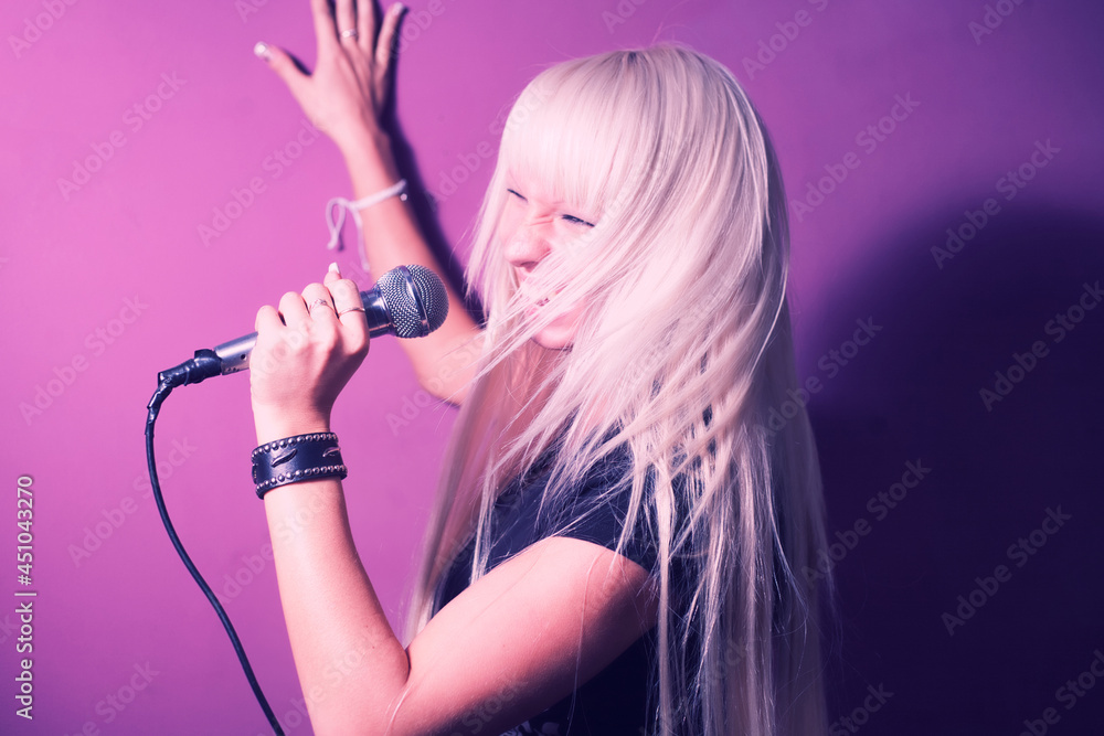Blonde woman in karaoke sing with microphone on pink backgound. She is in motion and her lond hair shaking around her head Blonde woman in karaoke sing with microphone on pink backgound. She is in motion and her lond hair shaking around her head