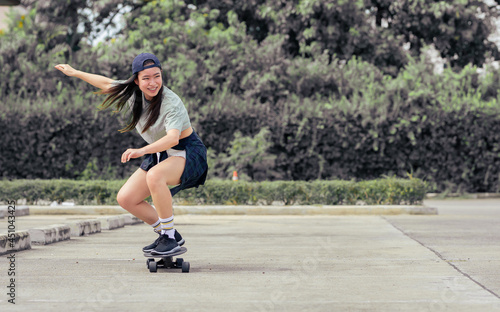 Portrait beautiful sportive Asian female skater wearing hipster shirt with shorts, smiling with happiness, standing on skateboard and playing outdoor with copy space. Activity and Adventure Concept.