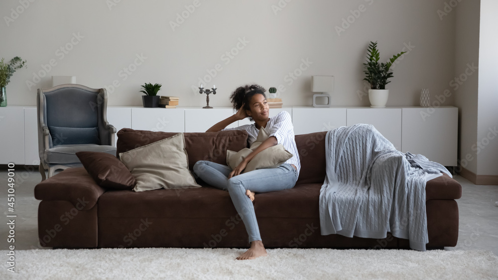 Happy calm millennial African American woman sit relax on sofa in living room look in distance dreaming. Smiling young mixed race female rest on couch at home breathe fresh ventilated condition air. Happy calm millennial African American woman sit relax on sofa in living room look in distance dreaming. Smiling young mixed race female rest on couch at home breathe fresh ventilated condition air.