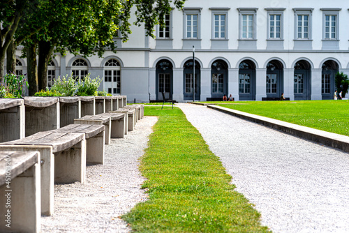 Benches and tables with flowers arranged in a row in the park, in the background a large building with arched windows.