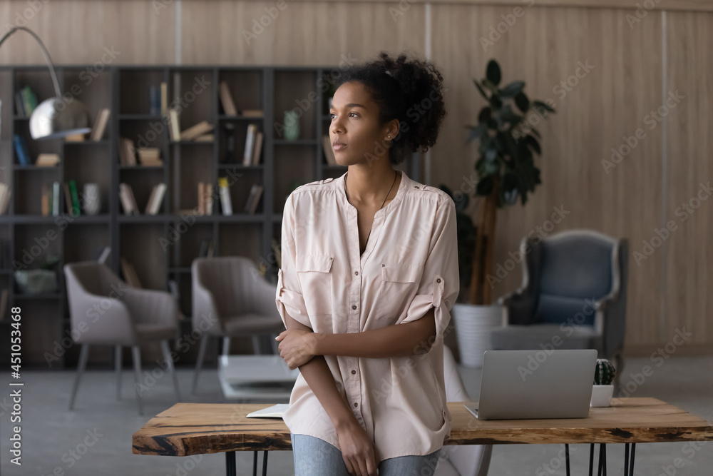 Pensive young African American woman at desk at home office look in distance thinking making plans. Thoughtful mixed race female lost in thoughts ponder visualize future perspectives opportunities.