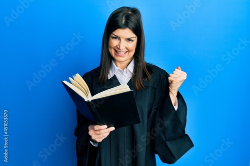 Young hispanic woman wearing judge uniform reading book screaming proud, celebrating victory and success very excited with raised arm