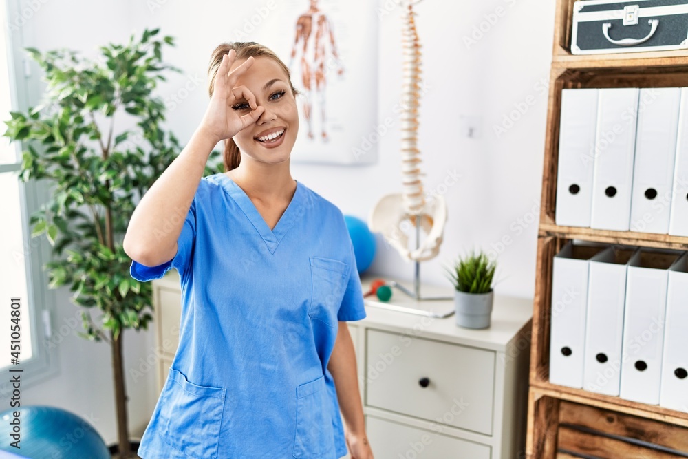 Young caucasian woman working at pain recovery clinic doing ok gesture with hand smiling, eye looking through fingers with happy face. Young caucasian woman working at pain recovery clinic doing ok gesture with hand smiling, eye looking through fingers with happy face.