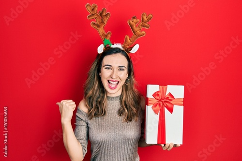 Young hispanic girl wearing deer christmas hat holding gift pointing thumb up to the side smiling happy with open mouth