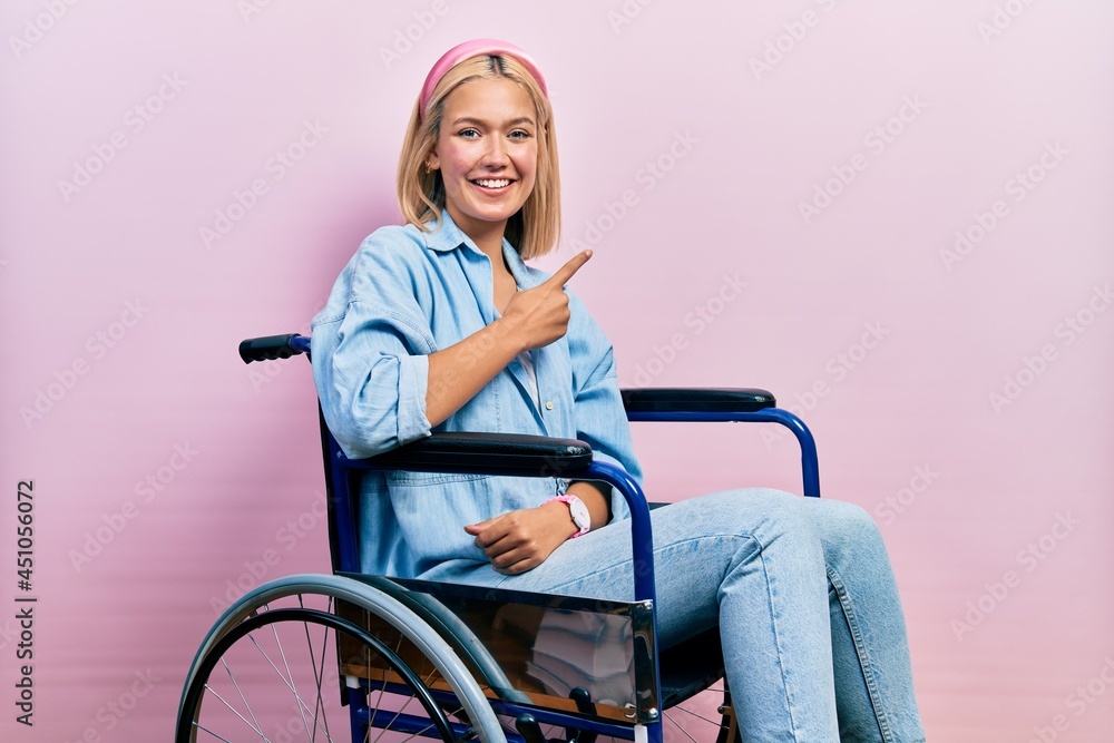 Beautiful blonde woman sitting on wheelchair cheerful with a smile on face pointing with hand and finger up to the side with happy and natural expression Beautiful blonde woman sitting on wheelchair cheerful with a smile on face pointing with hand and finger up to the side with happy and natural expression