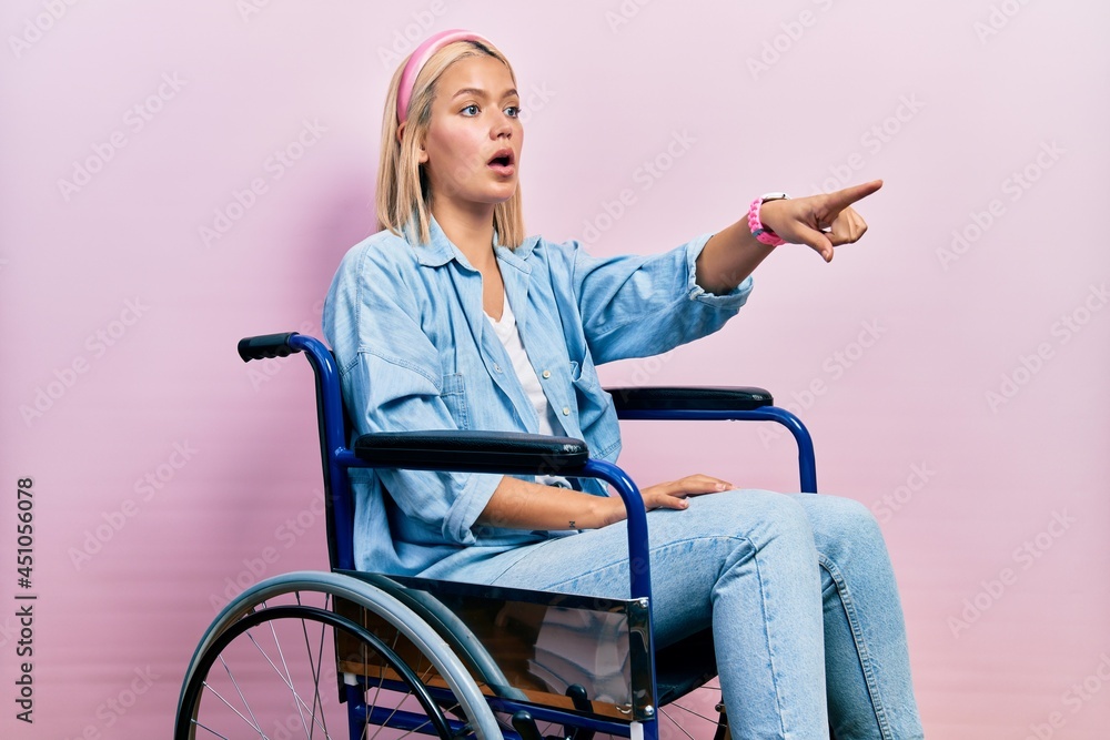 Beautiful blonde woman sitting on wheelchair pointing with finger surprised ahead, open mouth amazed expression, something on the front Beautiful blonde woman sitting on wheelchair pointing with finger surprised ahead, open mouth amazed expression, something on the front