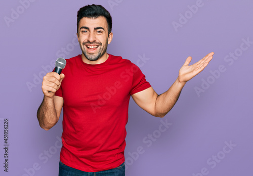 Hispanic man with beard singing song using microphone celebrating victory with happy smile and winner expression with raised hands