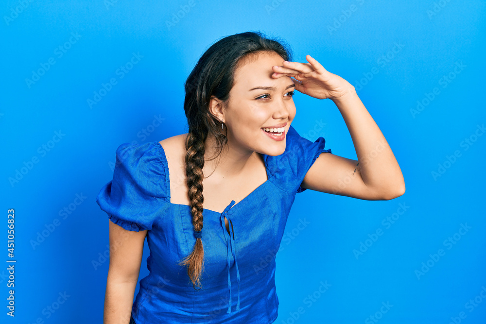 Young hispanic girl wearing casual clothes very happy and smiling looking far away with hand over head. searching concept. Young hispanic girl wearing casual clothes very happy and smiling looking far away with hand over head. searching concept.