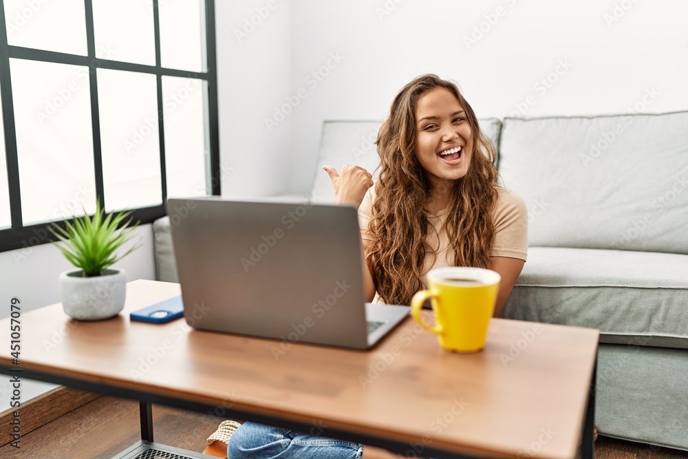 Beautiful hispanic woman using computer laptop at home smiling with happy face looking and pointing to the side with thumb up. Beautiful hispanic woman using computer laptop at home smiling with happy face looking and pointing to the side with thumb up.