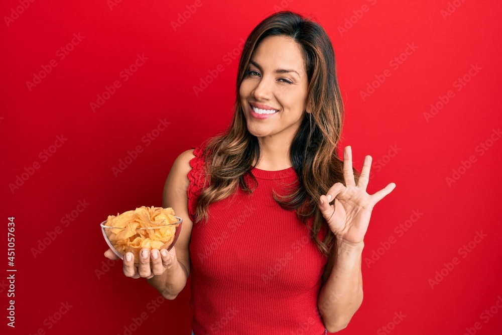 Young latin woman holding potato chip doing ok sign with fingers, smiling friendly gesturing excellent symbol Young latin woman holding potato chip doing ok sign with fingers, smiling friendly gesturing excellent symbol