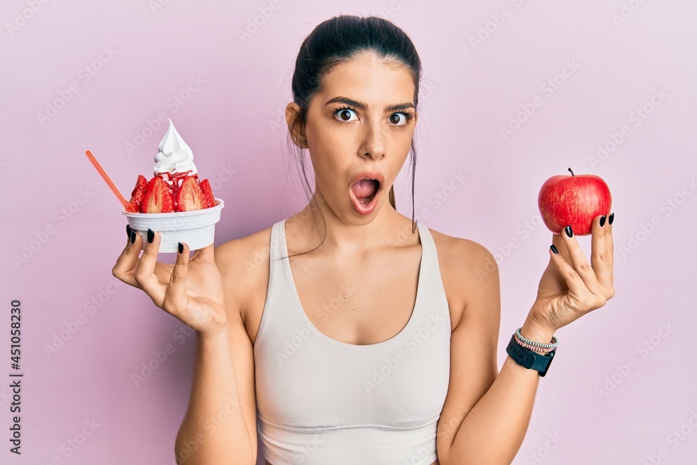 Young hispanic woman wearing sportswear holding apple and ice cream afraid and shocked with surprise and amazed expression, fear and excited face. Young hispanic woman wearing sportswear holding apple and ice cream afraid and shocked with surprise and amazed expression, fear and excited face.