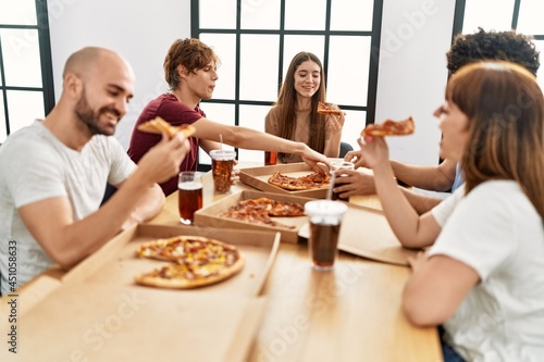 Group of young friends smiling happy eating italian pizza sitting on the table at home.