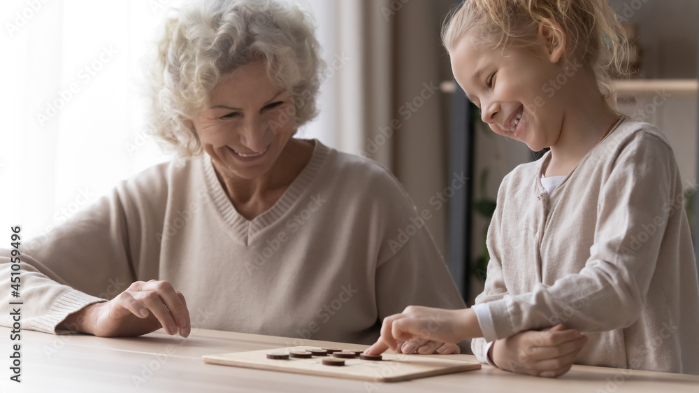 Smiling loving old grandmother and little granddaughter have fun play wooden board game together. Happy mature grandma and small grandchild feel playful with chess or draughts. Hobby concept. Smiling loving old grandmother and little granddaughter have fun play wooden board game together. Happy mature grandma and small grandchild feel playful with chess or draughts. Hobby concept.