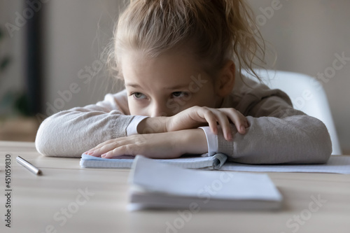 Sad little Caucasian girl child sit on table at home distracted from studying lack motivation doing homework. Unhappy small kid feel lazy bored preparing task assignment. Boredom, education concept. photo