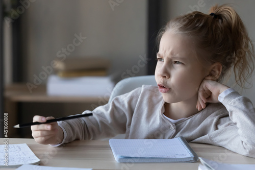 Unhappy little Caucasian girl kid sit at desk feel lazy unmotivated doing homework distressed or bothered with assignment. Upset child lack motivation writing preparing home task. Education concept.