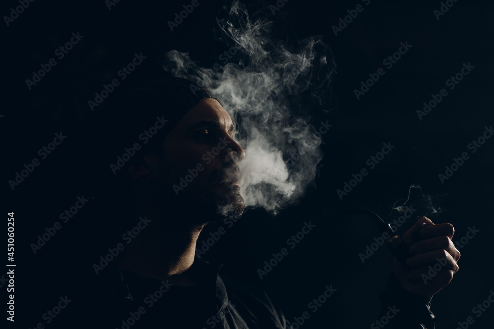 Man smokes smoking pipe against dark background. Back side lit profile portrait with smoke Man smokes smoking pipe against dark background. Back side lit profile portrait with smoke