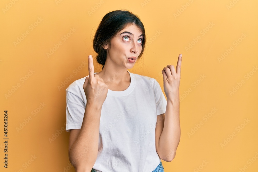Young caucasian woman wearing casual white t shirt amazed and surprised looking up and pointing with fingers and raised arms. Young caucasian woman wearing casual white t shirt amazed and surprised looking up and pointing with fingers and raised arms.