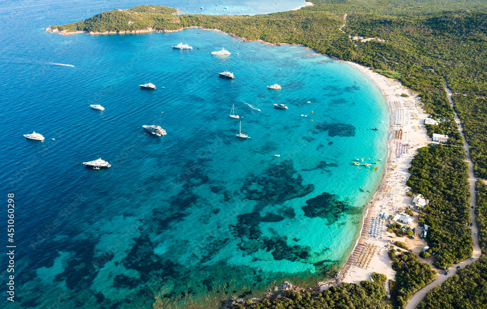 View from above, stunning aerial view of a green coastline with a white sand beach and and boats sailing on a turquoise water at sunset. Cala di volpe beach, Costa Smeralda, Sardinia, Italy.