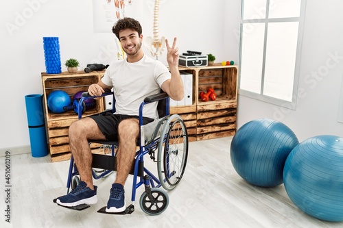 Hispanic man sitting on wheelchair at physiotherapy clinic smiling with happy face winking at the camera doing victory sign. number two.